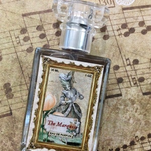 May include: A clear glass bottle of perfume with a gold-trimmed label. The label features a vintage illustration of a woman in a dress holding a flower. The text on the label reads "The Marquise Eau de Parfum 30 ML 1 OZ".