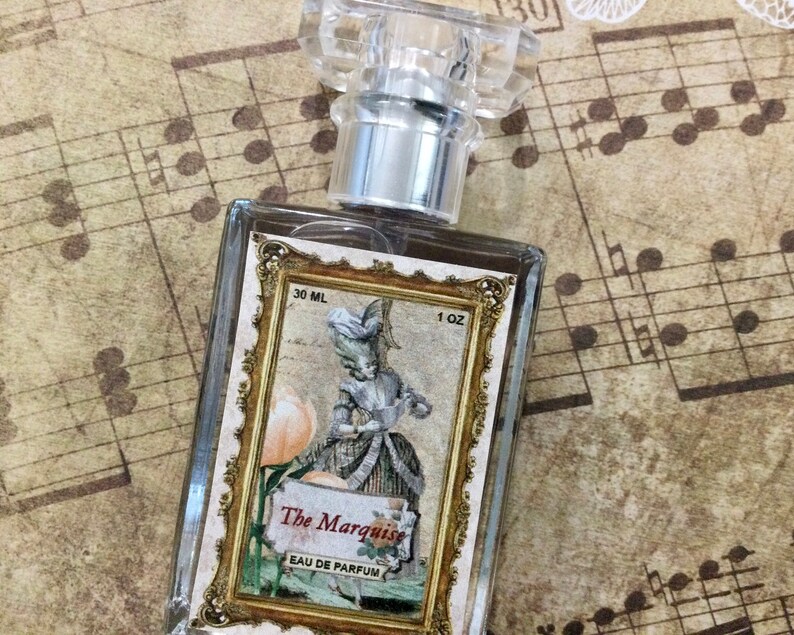 May include: A clear glass bottle of perfume with a gold-trimmed label. The label features a vintage illustration of a woman in a dress holding a flower. The text on the label reads "The Marquise Eau de Parfum 30 ML 1 OZ".