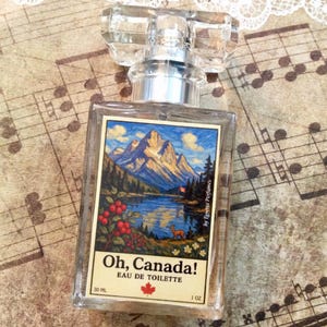May include: A clear glass perfume bottle with a rectangular label featuring a mountain landscape scene. The label reads "Oh, Canada! Eau de Toilette" with a maple leaf. The bottle contains 1 oz of perfume.