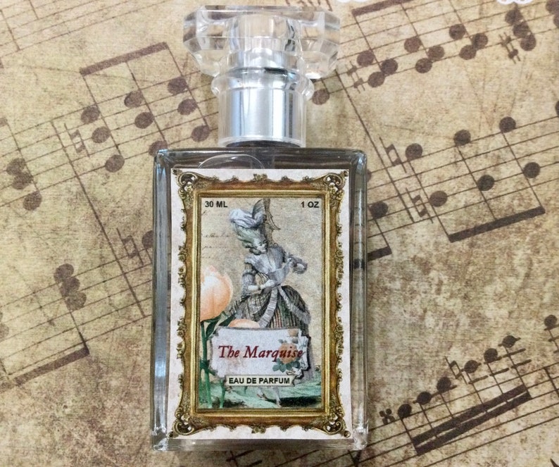 May include: A clear glass bottle of perfume with a silver cap. The bottle has a label with a gold frame and an illustration of a woman in a dress. The label reads "The Marquise Eau de Parfum" and "30 ML 1 OZ".