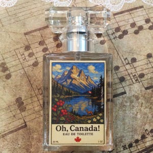 May include: A clear glass perfume bottle with a faceted crystal top. The label features a landscape painting of mountains, a lake, and a deer. The text "Oh, Canada! Eau de Toilette" is printed on the label. The bottle contains 1 fl oz (30 ml).