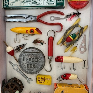 May include: A vintage wooden box filled with fishing lures, tackle, and tools. The box includes a red and yellow fishing lure, a red and white fishing lure, a leader box, a pair of pliers, a fishing line dressing tin, and a South Bend fishing lure box.