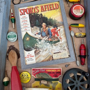 May include: A vintage wooden box filled with fishing supplies, including a magazine titled "Sports Afield", a fishing reel, lures, and a wooden spoon. The box is lined with burlap fabric.