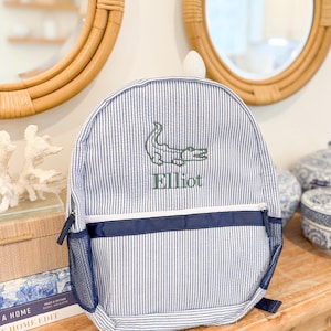May include: Blue and white striped backpack with a green embroidered alligator and the name "Elliot". The backpack has a navy blue accent stripe and mesh side pocket. A stylish and practical accessory for everyday use.