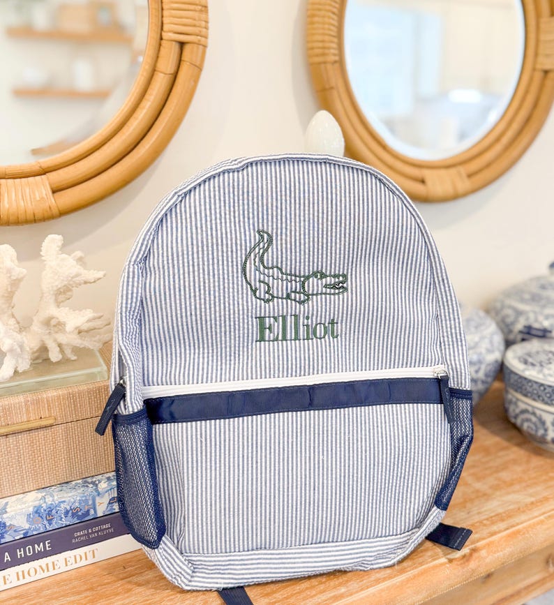 May include: Blue and white striped backpack with a green embroidered alligator and the name "Elliot". The backpack has a navy blue accent stripe and mesh side pocket. A stylish and practical accessory for everyday use.