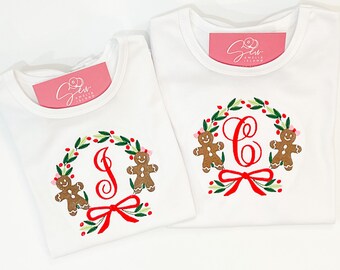 May include: Two white t-shirts with a Christmas wreath design. Each wreath features a red initial, gingerbread men, red berries, and a red bow. The shirts have a tag that says "Sew Amelia Island".