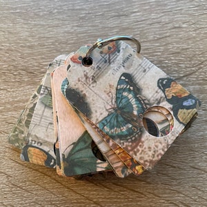 May include: A stack of six decorative tags with a butterfly design and musical notes. The tags are made of paper and have a silver ring attached to the top.