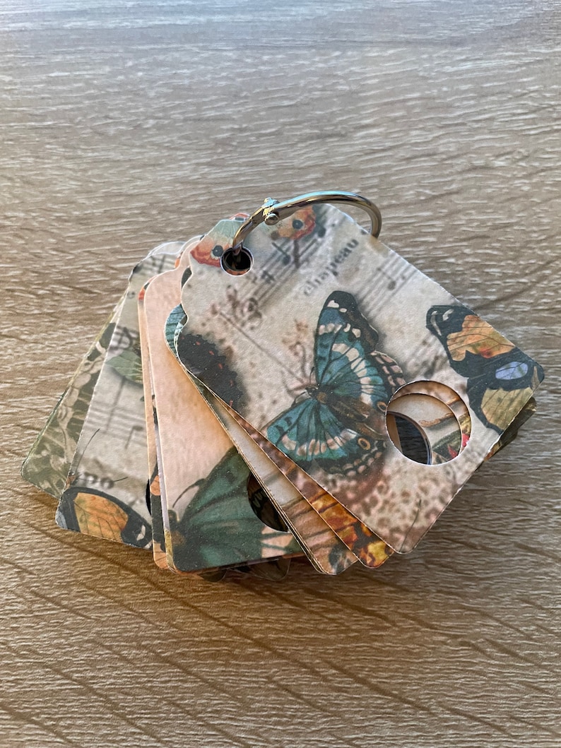 May include: A stack of six decorative tags with a butterfly design and musical notes. The tags are made of paper and have a silver ring attached to the top.