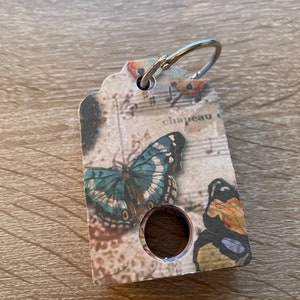 May include: A key chain with a silver ring and a tag featuring a butterfly design. The tag is decorated with a vintage music sheet background and the word "chapeau" is visible.