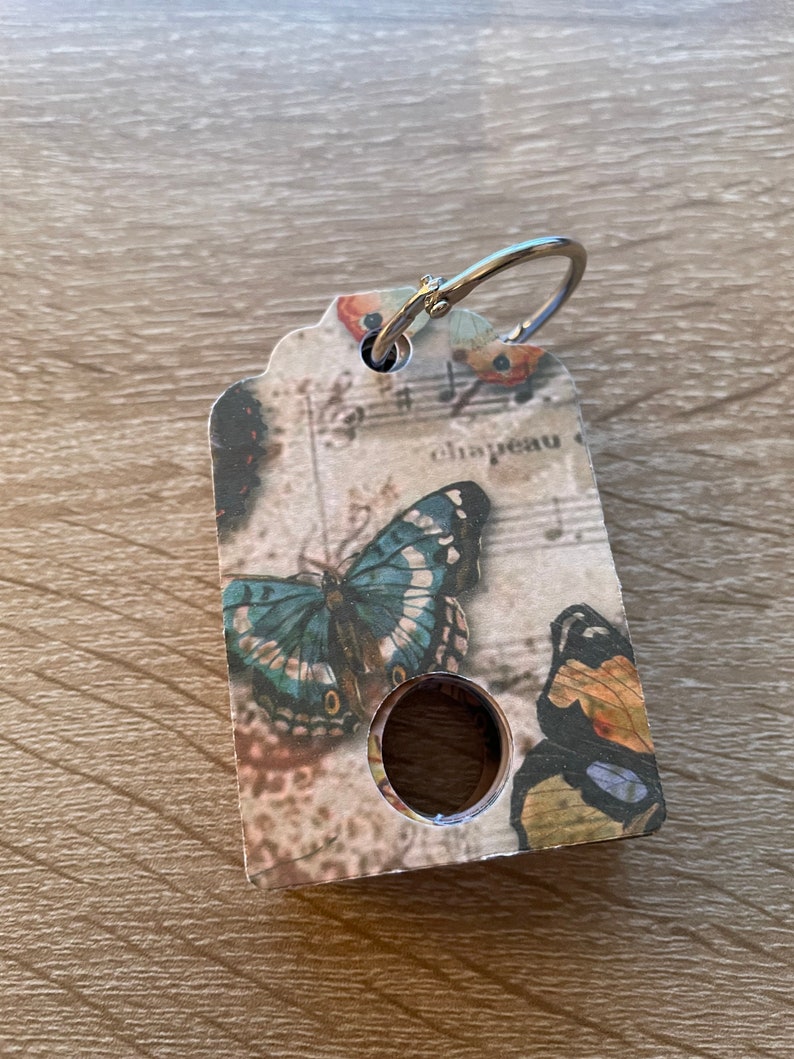 May include: A key chain with a silver ring and a tag featuring a butterfly design. The tag is decorated with a vintage music sheet background and the word "chapeau" is visible.
