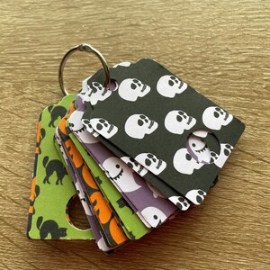 May include: A set of Halloween-themed decorative tags on a silver metal ring. The tags display patterns such as white skulls on a black background, ghosts, and black cats on green and orange backgrounds. The tags are made of paper and have a circular hole.