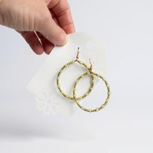 May include: A pair of gold-tone hoop earrings with a beaded design. The earrings feature small, light green and gold beads strung together to form a circular shape. The earrings are displayed on a white tag with a decorative cut-out.