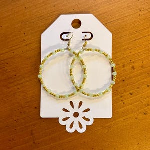 May include: A pair of gold and light green beaded hoop earrings. The earrings are displayed on a white tag with a flower-shaped cutout at the bottom. The earrings have a gold-colored metal hook.