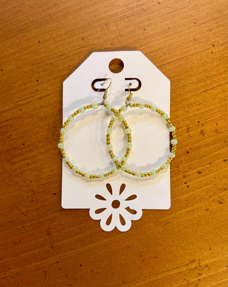 May include: A pair of gold and light green beaded hoop earrings. The earrings are displayed on a white tag with a flower-shaped cutout at the bottom. The earrings have a gold-colored metal hook.