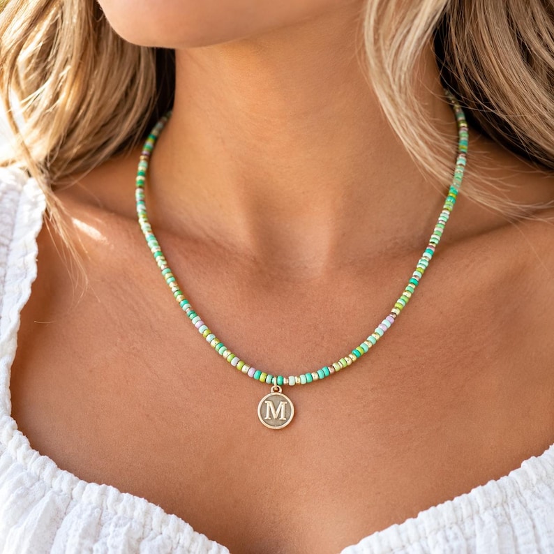 May include: A beaded necklace featuring a strand of small, multi-colored glass beads in shades of green, yellow, white, and blue. The necklace includes a circular gold-toned pendant centered at the bottom, which features the capital letter M.