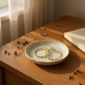 May include: A pair of gold hoop earrings with light green and gold beads rests on a small, off-white ceramic dish. The earrings are displayed on a wooden surface, with a book and dried lavender sprigs nearby.