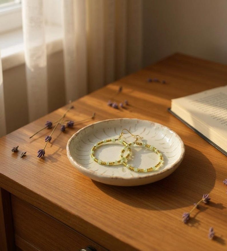 May include: A pair of gold hoop earrings with light green and gold beads rests on a small, off-white ceramic dish. The earrings are displayed on a wooden surface, with a book and dried lavender sprigs nearby.