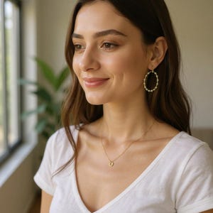 May include: A woman wearing gold-colored jewelry, including a necklace with a small pendant and oval hoop earrings. The earrings and necklace appear to be adorned with small, white beads. She is wearing a white v-neck shirt.