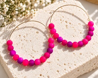 May include: A pair of hoop earrings featuring thin gold-toned metal wires adorned with a series of matte spherical beads in alternating shades of vibrant hot pink and deep purple, arranged along the lower curve of each circular earring frame.