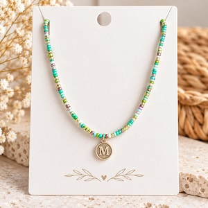 May include: A beaded necklace featuring small, multi-colored glass beads in shades of turquoise, green, gold, and white. The necklace includes a circular gold-toned metal pendant embossed with the capital letter M, centered on a white display card.