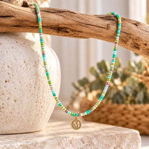 May include: A beaded necklace featuring a repeating pattern of small turquoise, lime green, metallic gold, and light purple seed beads. A circular gold-toned metal charm with the embossed capital letter M hangs from the center of the necklace.
