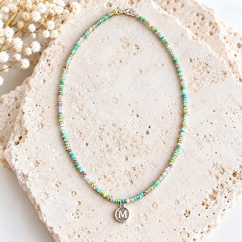May include: A beaded necklace featuring small, multi-colored glass beads in shades of green, turquoise, white, and metallic silver. The necklace includes a circular silver-toned charm with the letter M engraved in the center and a lobster clasp.