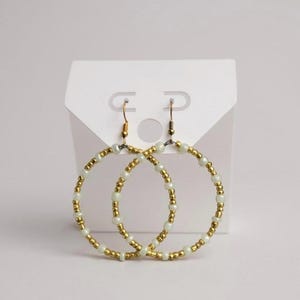 May include: A pair of gold-colored hoop earrings adorned with small, round, off-white beads. The earrings are displayed on a white earring card, showcasing their design and craftsmanship. The earrings have a simple, elegant design.