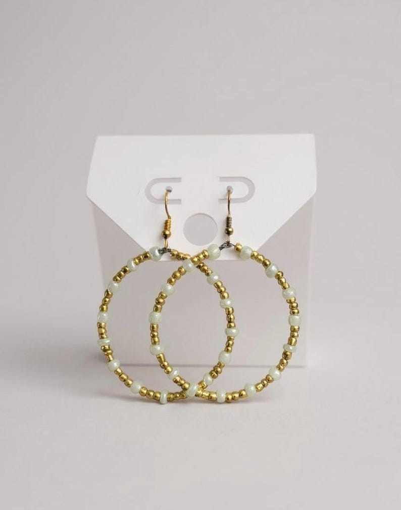 May include: A pair of gold-colored hoop earrings adorned with small, round, off-white beads. The earrings are displayed on a white earring card, showcasing their design and craftsmanship. The earrings have a simple, elegant design.