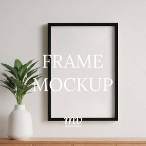 Puede incluir: Una maqueta de marco rectangular negro con el texto "FRAME MOCKUP" en blanco. Un jarrón blanco con una planta de hojas verdes está sobre una superficie de madera. El fondo es una pared de color beige claro.