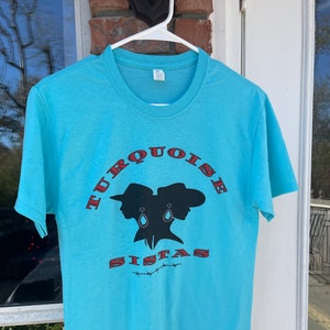 May include: A turquoise blue t-shirt with a red and black graphic of two women wearing cowboy hats and the text "TURQUOISE SISTAS".