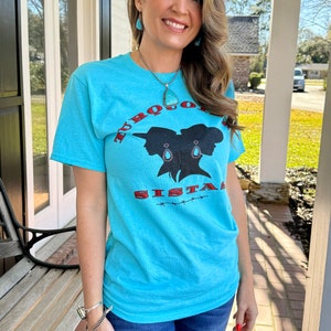 May include: A turquoise blue t-shirt with a graphic of two women's silhouettes wearing turquoise earrings and hats. The text "TURQUOISE SISTAS" is printed in red and black on the shirt.