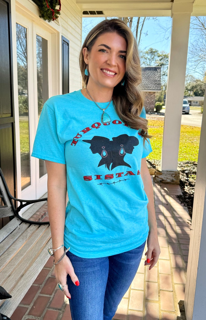 May include: A turquoise blue t-shirt with a graphic of two women's silhouettes wearing turquoise earrings and hats. The text "TURQUOISE SISTAS" is printed in red and black on the shirt.