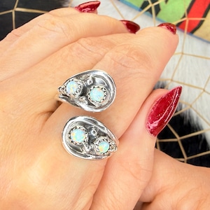 May include: Two silver rings, each featuring a unique design with two oval-shaped, light blue opal stones. The rings are set in a detailed, textured setting, showcasing a vintage or handcrafted aesthetic. The rings are on a hand with red nail polish.