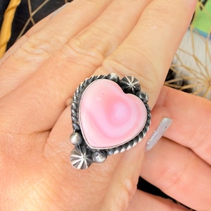 May include: A silver-toned ring featuring a large, heart-shaped pink stone. The stone is set in a decorative frame with a twisted rope design and accented with small, star-shaped details. The ring is shown on a hand.