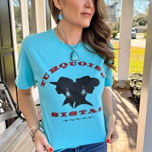 May include: A turquoise blue t-shirt featuring a graphic of two women's silhouettes wearing cowboy hats and turquoise earrings. The text "TURQUOISE SISTAS" is printed in red and black on the shirt.
