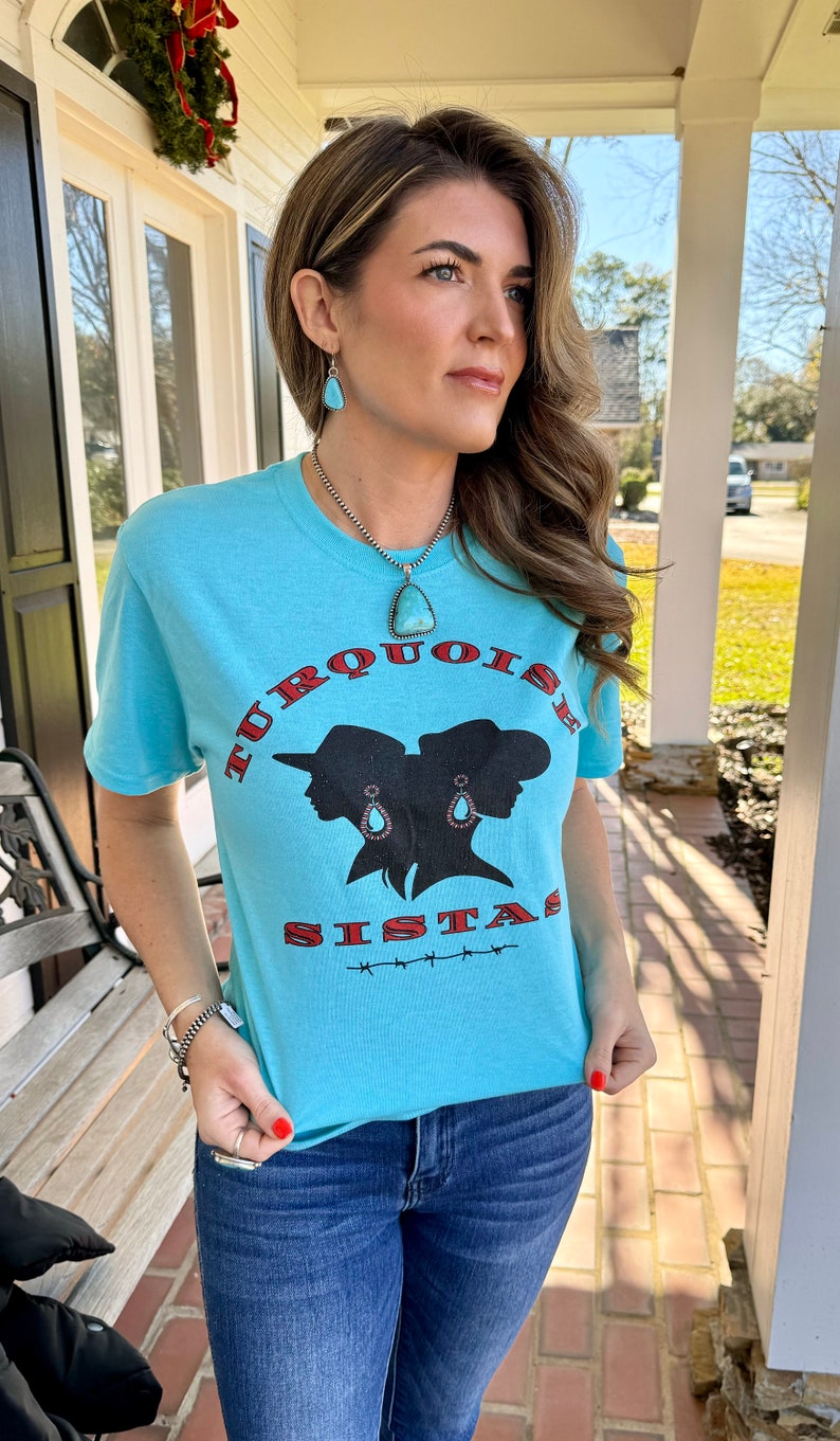 May include: A turquoise blue t-shirt featuring a graphic of two women's silhouettes wearing cowboy hats and turquoise earrings. The text "TURQUOISE SISTAS" is printed in red and black on the shirt.