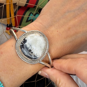 May include: A silver cuff bracelet featuring a large, oval-shaped stone with a marbled white and black pattern. The stone is set in a smooth silver bezel surrounded by a twisted rope-style silver border and a polished silver frame.
