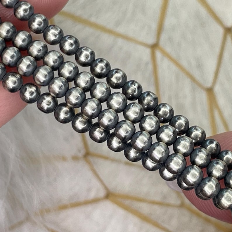 Peut inclure: Gros plan sur plusieurs brins de petites perles rondes gris fonc&eacute;. Les perles ont un &eacute;clat m&eacute;tallique et sont enfil&eacute;es ensemble, cr&eacute;ant une surface textur&eacute;e. Le fond est de couleur claire.