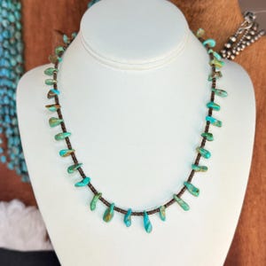 May include: A turquoise stone necklace displayed on a white mannequin. The necklace features teardrop-shaped turquoise stones strung on a dark brown cord. The stones vary in shades of blue and green, creating a natural, earthy aesthetic.