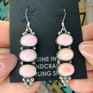 May include: A pair of dangle earrings featuring three vertically aligned oval pink stones set in oxidized sterling silver. The earrings have a decorative beaded metal base and silver ear wires, displayed on a black card with visible text.