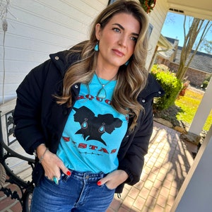 May include: A woman wearing a black puffer jacket, blue jeans, and a turquoise blue t-shirt with a graphic of two women's silhouettes and the text "BOUNCE SISTAS". She is wearing turquoise earrings and a turquoise necklace.