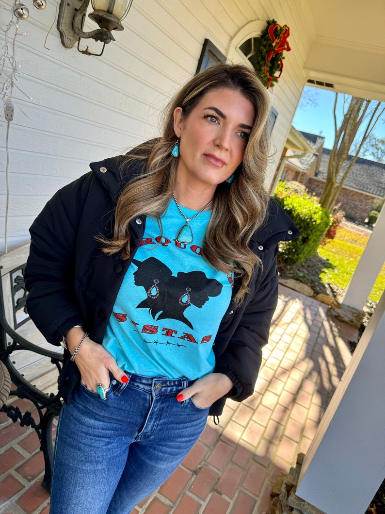 May include: A woman wearing a black puffer jacket, blue jeans, and a turquoise blue t-shirt with a graphic of two women's silhouettes and the text "BOUNCE SISTAS". She is wearing turquoise earrings and a turquoise necklace.