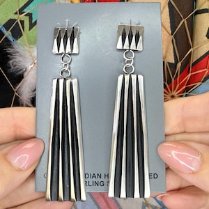 May include: A pair of long, dangling earrings featuring a silver-toned top square component with three vertical ridges, connected by a small jump ring to a larger, fan-shaped silver-toned base with deep black vertical grooves and a polished finish.