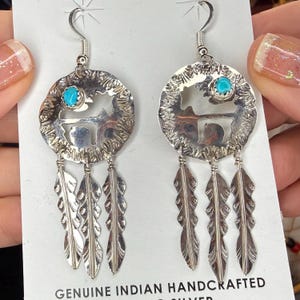 May include: Silver dangle earrings with a turquoise stone and a cut-out design of a horse and landscape. The earrings have three dangling feather charms. The text "GENUINE INDIAN HANDCRAFTED SILVER" is visible.