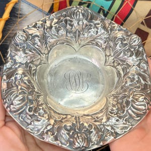 May include: Ornate silver dish with a floral and leaf design around the rim. The center of the dish features an engraved monogram. The dish has a scalloped edge and is held in a person's hands.