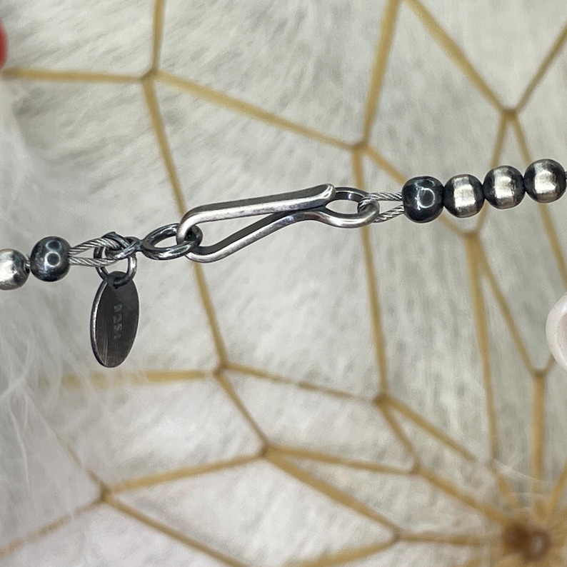 Peut inclure: Gros plan sur un fermoir de collier argent&eacute; avec une petite &eacute;tiquette ronde. Le collier pr&eacute;sente un m&eacute;lange de perles argent&eacute;es et gris fonc&eacute;. Le fermoir est de type crochet et &oelig;illet, et l'&eacute;tiquette porte une inscription.