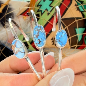 May include: Three silver cuff bracelets, each featuring a single oval-shaped light blue stone with dark brown matrix veining. The bracelets have a polished metallic finish and an open-ended design, showcasing the smooth texture of the stones.