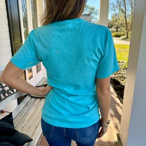 May include: A woman wearing a light blue t-shirt and blue jeans. The t-shirt is a simple design with no visible graphics or text.