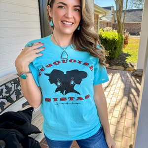 May include: A turquoise blue t-shirt with a graphic of two women wearing turquoise earrings and the text "Turquoise Sistahs" in red.