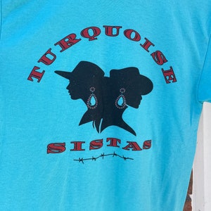 May include: A turquoise blue t-shirt featuring a black silhouette of two women wearing cowboy hats and earrings. The text "TURQUOISE SISTAS" is printed in red on the shirt.